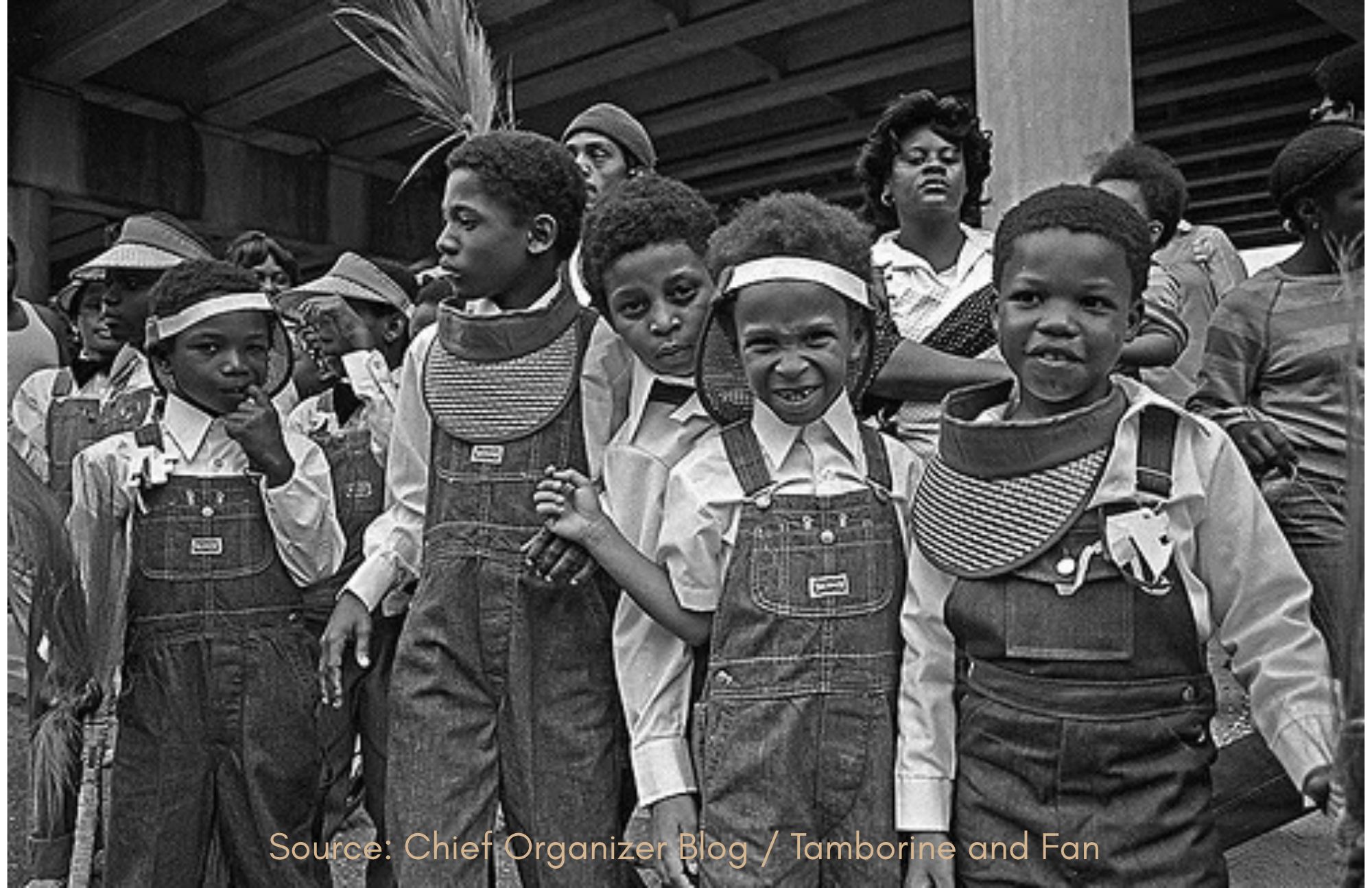 Black-and-white photograph of young boys in matching overalls and visors gathered with community members beneath an overpass in New Orleans. Source: Chief Organizer Blog / Tamborine and Fan