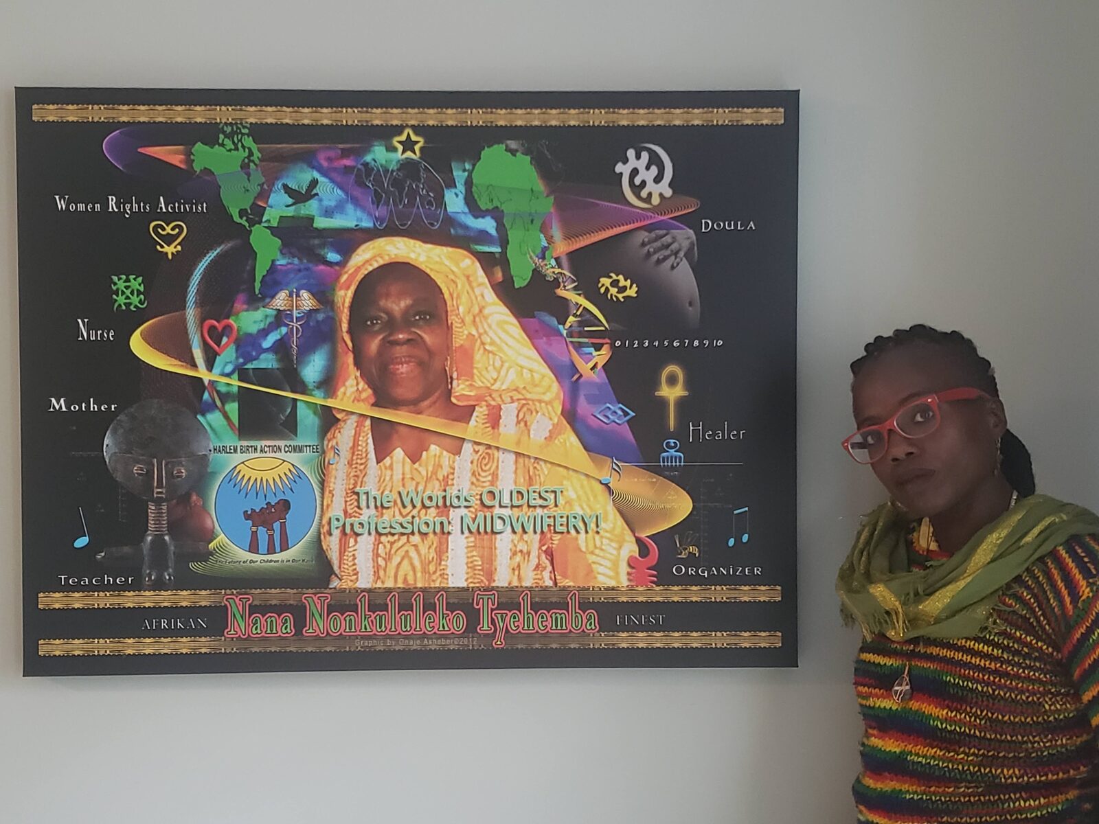 A woman in red glasses and a striped knit sweater stands beside a large framed tribute poster to Nana Nonkululeko Tyehemba, honoring her as a midwife, doula, nurse, healer, teacher, organizer and founder of the Harlem Birth Action Committee