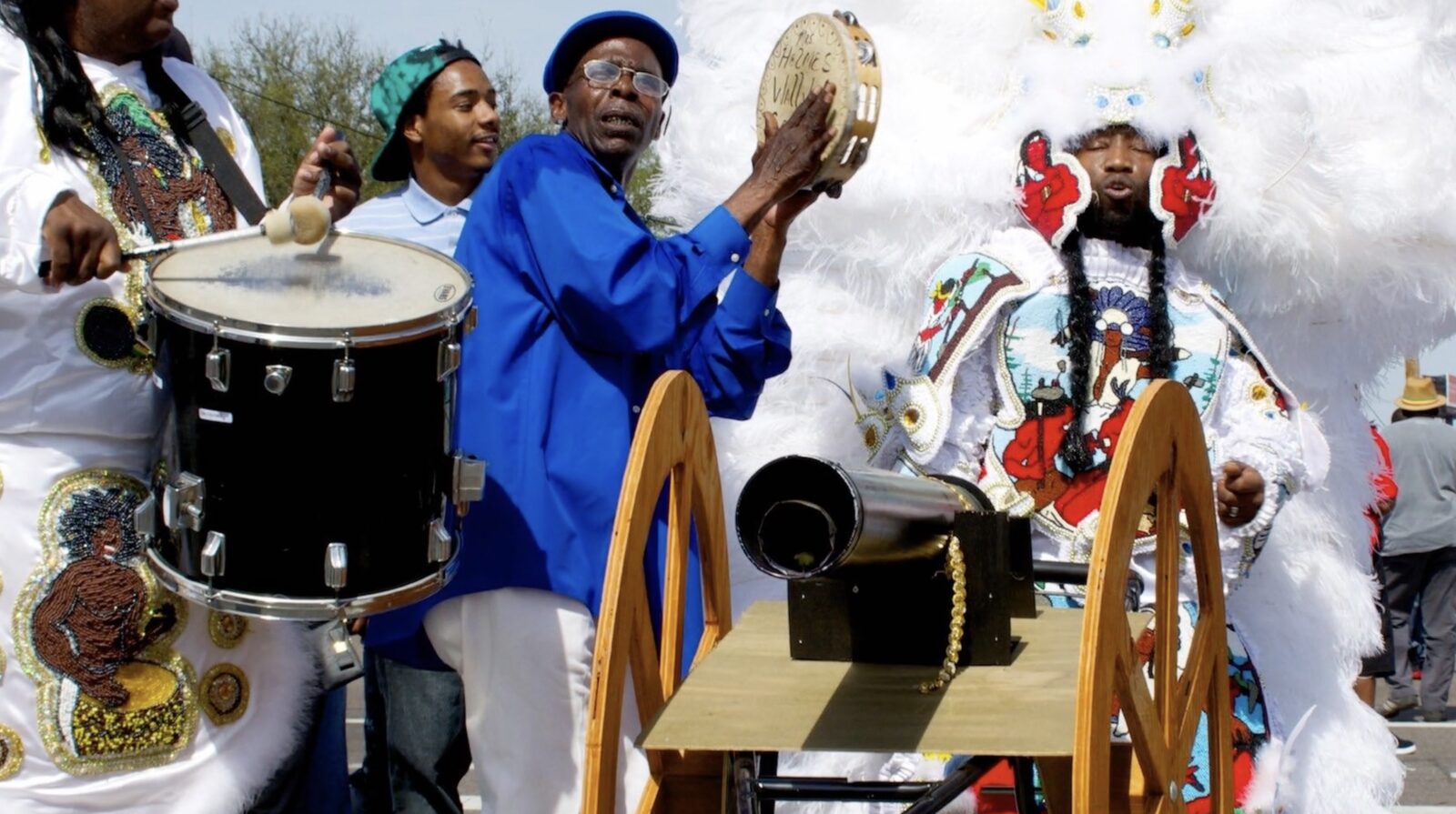 Still from The Needle and The Thread: drummers and a tambourine player beside a Black Indian in a white feathered suit